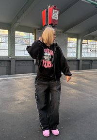 Crna oversized dukserica s ukrasima od zakovica, majica s grafikom lica, crne traperice ukrašene cvjetnim motivima i ružičaste papuče.