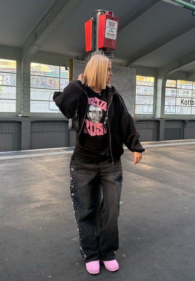 Sudadera negra oversize con acentos de tachuelas, camiseta gráfica con una cara, jeans negros adornados con embellishments florales y pantuflas rosas.