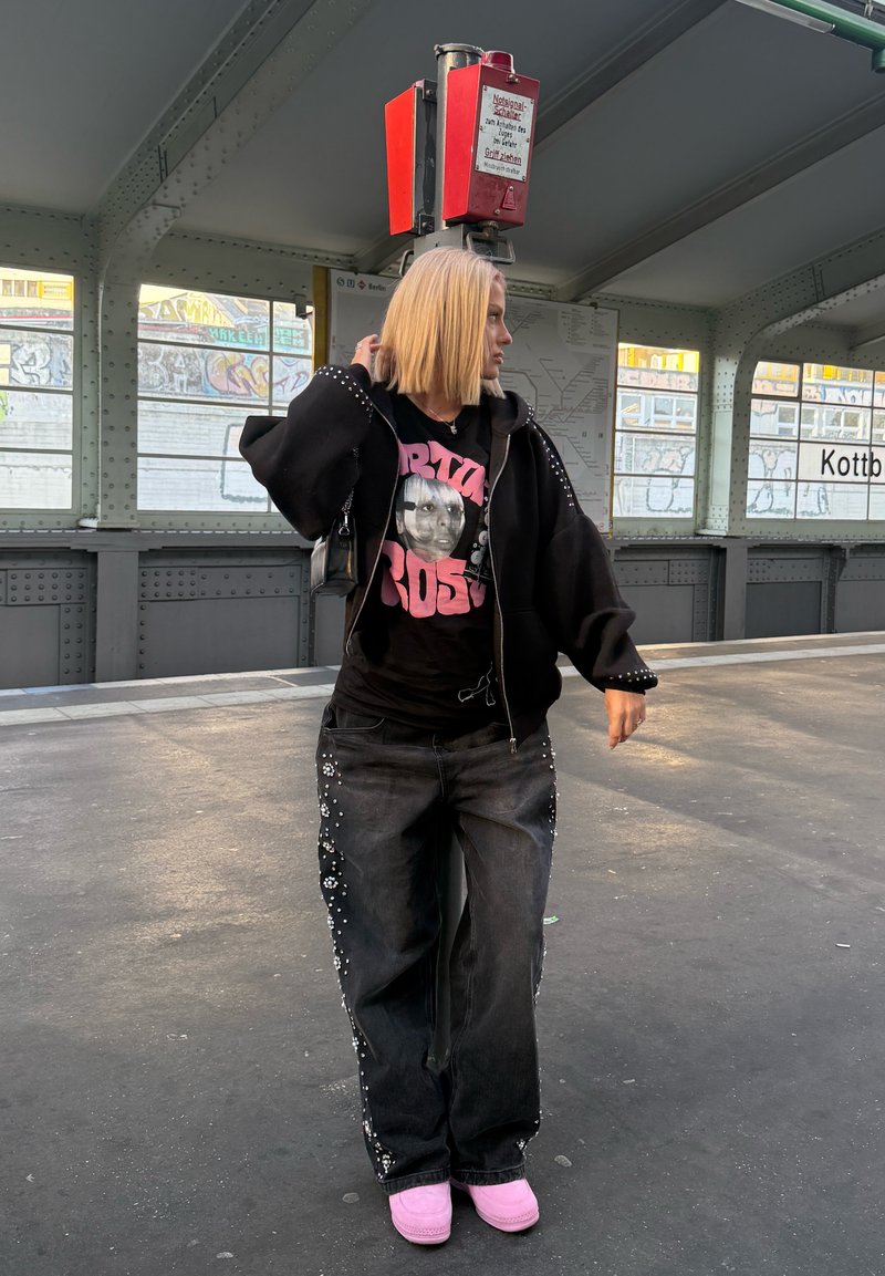 Sweatshirt oversized preto com detalhes em tachas, t-shirt gráfica com um rosto, jeans pretos adornados com embellishments florais, e chinelos cor-de-rosa.