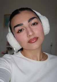 Weiße, flauschige Ohrenschützer umrahmen das Gesicht und kontrastieren mit der dunklen Haarfarbe. Das Model trägt ein cremefarbenes Oberteil, das glänzende Lippen und ein leichtes Makeup zur Geltung bringt.