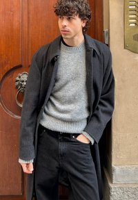 Manteau noir sur un pull gris avec un ourlet côtelé, associé à un jean noir. La texture semble douce et le manteau a une coupe ajustée.