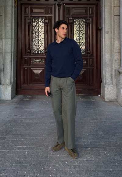 Joven vestido con un suéter azul marino y pantalones grises, de pie con una mano en el bolsillo frente a unas grandes puertas dobles de madera ornamentadas.