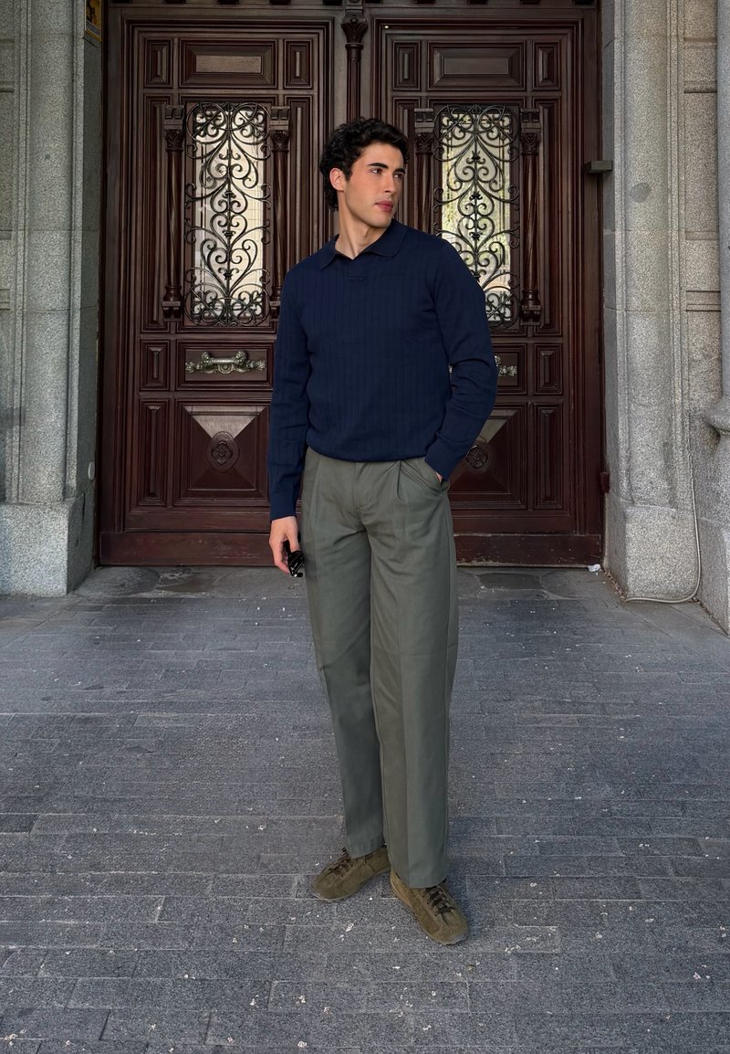 Jeune homme portant un pull bleu marine et un pantalon gris, debout avec une main dans la poche devant de grandes portes doubles en bois ornées.