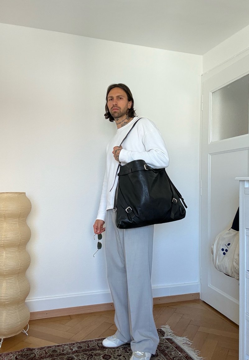 Homme debout à l'intérieur portant une chemise blanche à manches longues et un pantalon gris, tenant des lunettes de soleil et portant un grand sac à épaule en cuir noir.