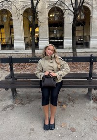 Beige kaksirivinen takki, mustat lyhyet housut ja mustat tasapäiset kengät. Kädet ruskeassa käsilaukussa, istumassa penkillä syksyn lehtien ympäröimänä.