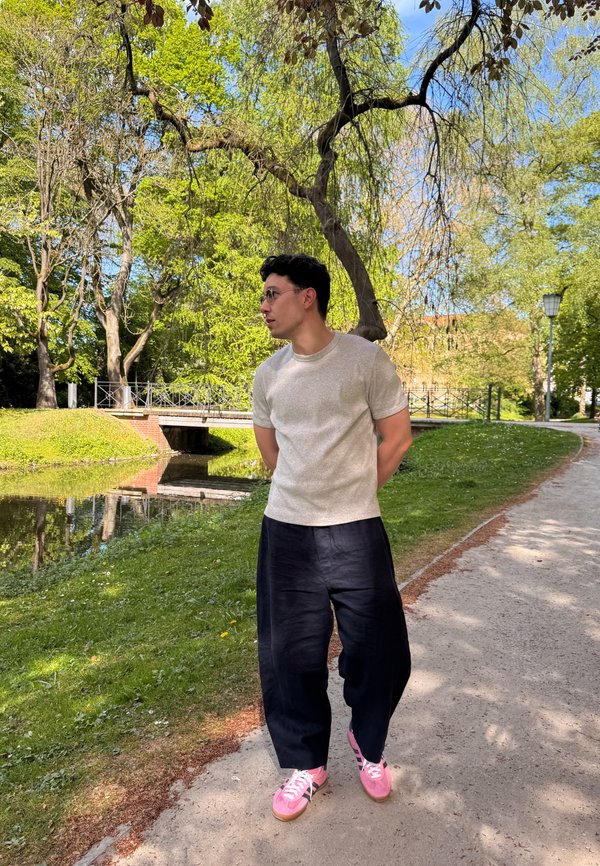 Man met een bril, wit t-shirt, zwarte broek en roze sneakers die langs een parkpad loopt naast een kleine waterloop en groene bomen.