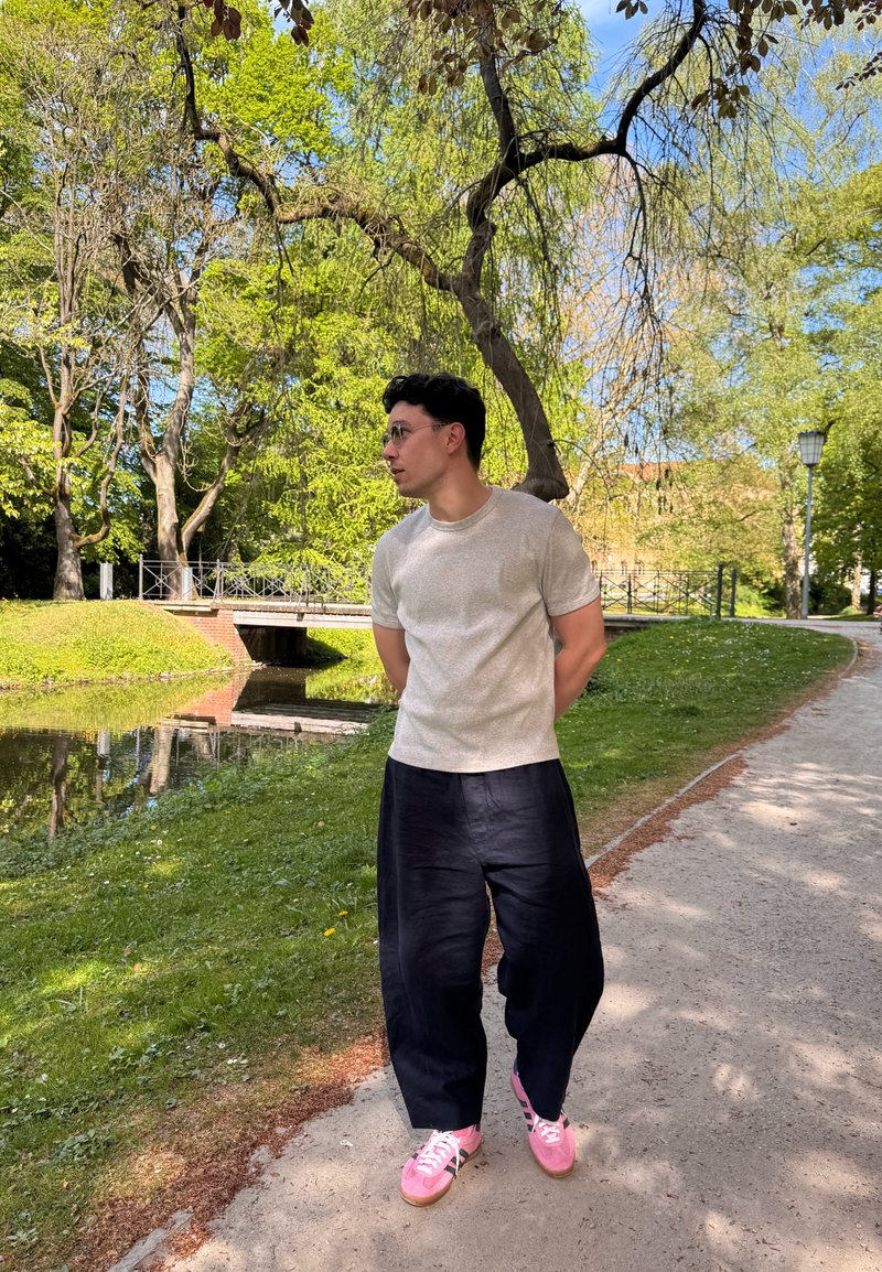 Homme portant des lunettes, un t-shirt blanc, un pantalon noir et des baskets roses marchant le long d'un chemin de parc près d'un petit cours d'eau et d'arbres verts.