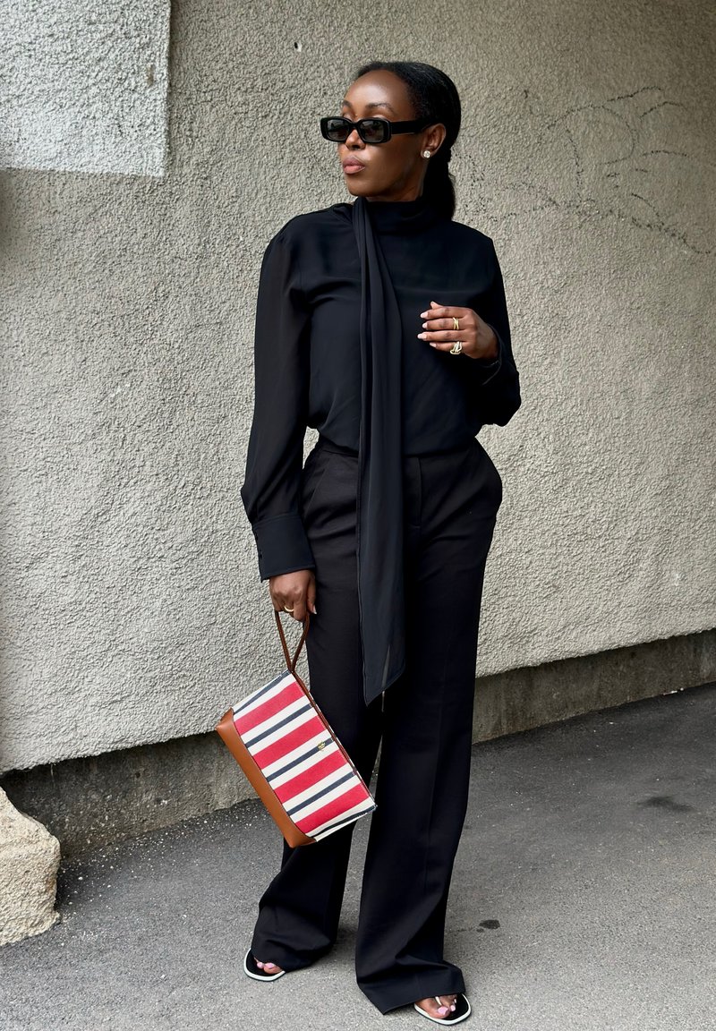 Woman wearing black outfit and sunglasses holding a striped red, white, and blue handbag standing against a textured wall.