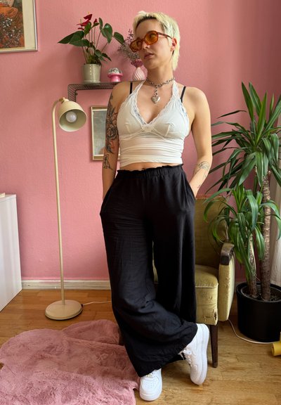 Top de cultivo blanco con encaje, pantalones anchos negros y zapatillas blancas. Las gafas de sol naranjas y el collar plateado realzan el conjunto. Fondo de pared rosa.