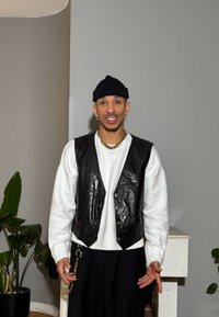 Gilet in pelle nera sopra un maglione bianco, pantaloni neri e gioielli dorati. Sullo sfondo una pianta e una parete grigia semplice.