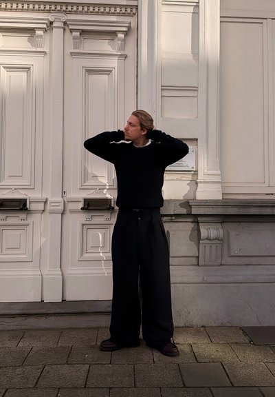 Hombre con cabello rubio, suéter negro y pantalones anchos de pie en una calle pavimentada, con las manos detrás de la cabeza, frente a unas grandes puertas blancas ornamentadas.