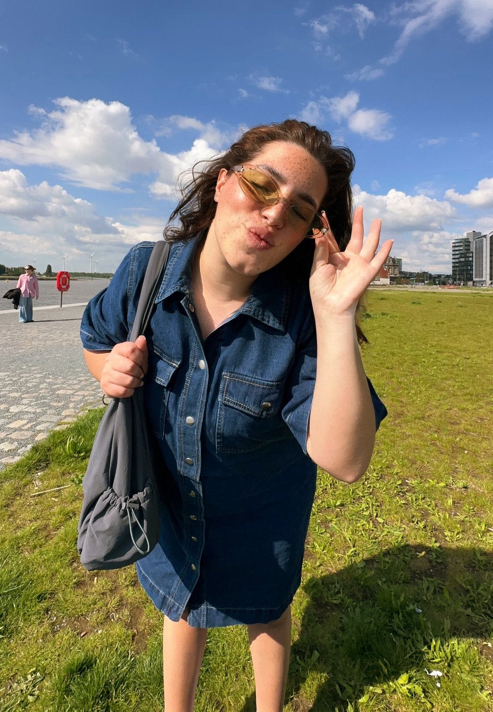 Denim πουκάμισο φόρεμα σε σκούρο μπλε με κουμπιά μπροστά και κοντά μανίκια. Φοράει γυαλιά ηλίου με κίτρινη απόχρωση, κρατώντας μια γκρι τσάντα με κορδόνι. Φόντο με πράσινο γρασίδι.