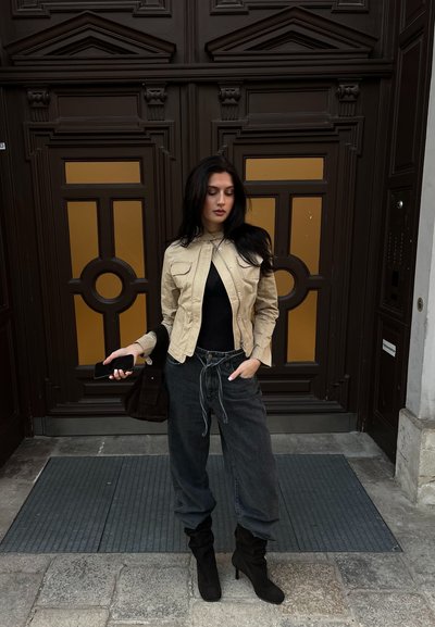 Chaqueta beige de corte cropped con bolsillos frontales, top negro, pantalones grises holgados y botas de tacón negras. Sosteniendo un teléfono y un bolso oscuro.