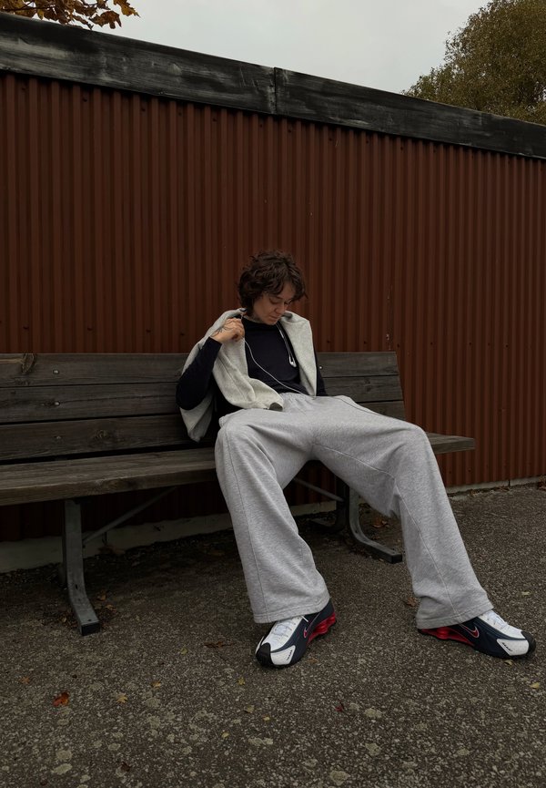 Conjunto casual com um hoodie sem mangas cinza sobre uma camisa de manga longa preta e calças de moletom cinzas largas, combinadas com ténis pretos e vermelhos.