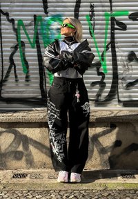 Črne široke hlače z belimi grafičnimi vzorci, oversized črna jakna, mrežasta bluza, zelena sončna očala in roza superge.