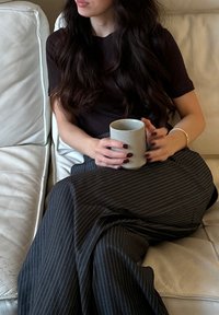 Una persona con una maglia scura tiene in mano una tazza di ceramica cilindrica. Indossa una gonna a righe bianche e nere, seduta su un divano di pelle chiara.