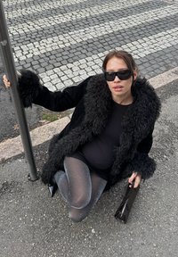 Femme enceinte en manteau de fourrure noir, collants scintillants et lunettes de soleil, assise sur le trottoir, tenant un poteau et un sac à main noir près d'un passage pour piétons.