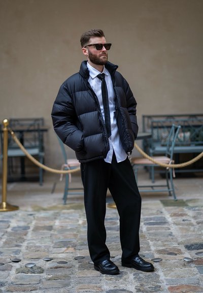 Chaqueta de plumas negra, camisa blanca, corbata negra y pantalones negros; combinados con mocasines negros y gafas de sol oscuras. Atuendo urbano casual.