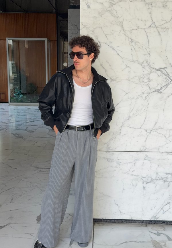Hombre con chaqueta de cuero negra, camiseta blanca sin mangas, pantalones grises de rayas y gafas de sol, de pie contra una pared de mármol blanco en el interior.