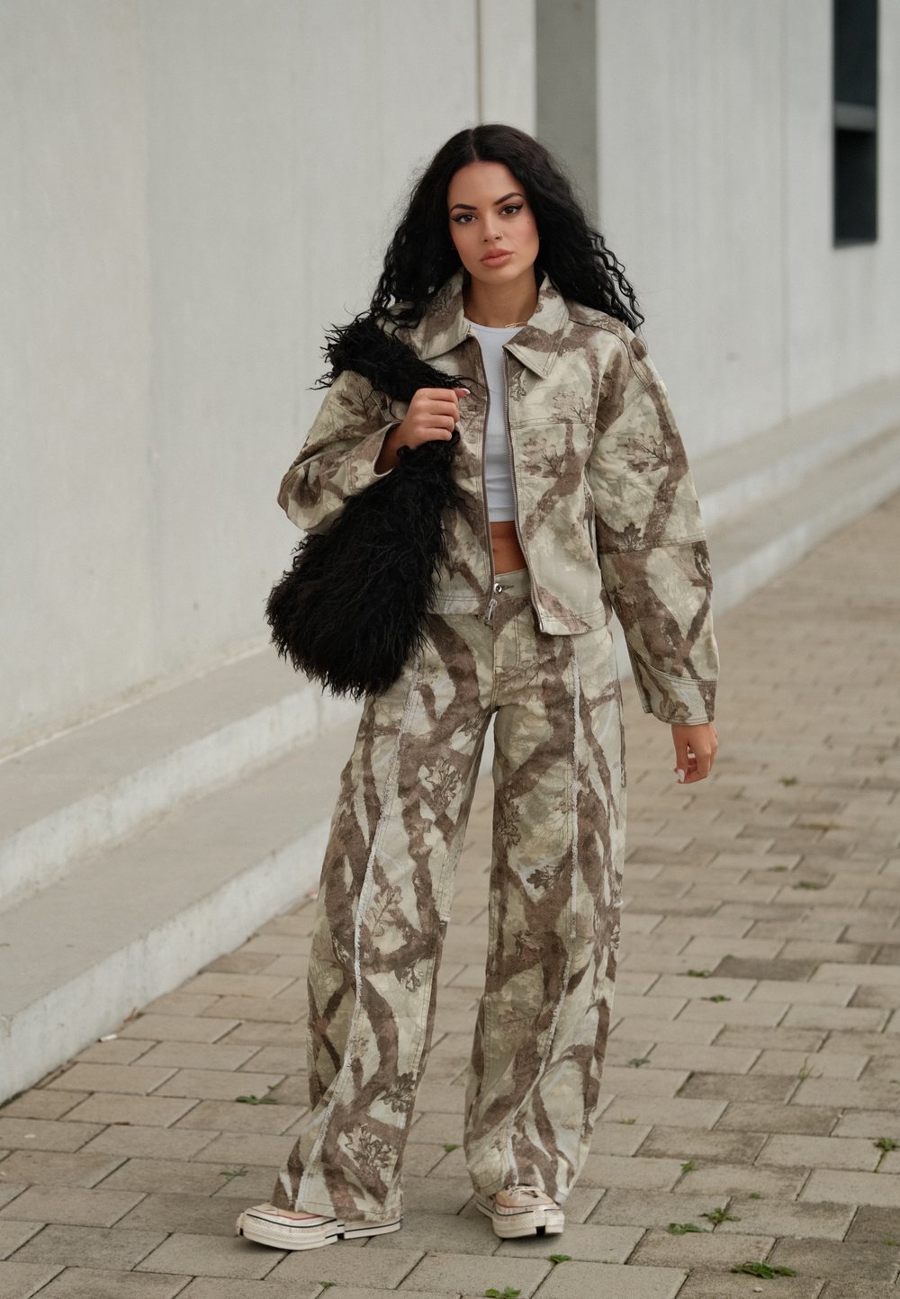 Kamouflage-mönstrad jacka och vida byxor i beige och brunt, kombinerat med en fluffig svart axelväska och vita sneakers.