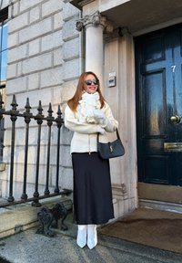 Femme portant des lunettes de soleil avec une écharpe blanche, des gants, une veste, une jupe noire et des bottes blanches, souriante, debout près de l'entrée d'un bâtiment en pierre avec une porte noire et une rambarde en fer.