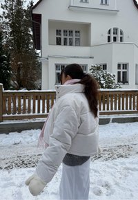 Άτομο με λευκό μπουφάν, ροζ κασκόλ και γάντια περπατά σε χιονισμένο μονοπάτι μπροστά από ένα λευκό σπίτι με ξύλινο φράχτη.