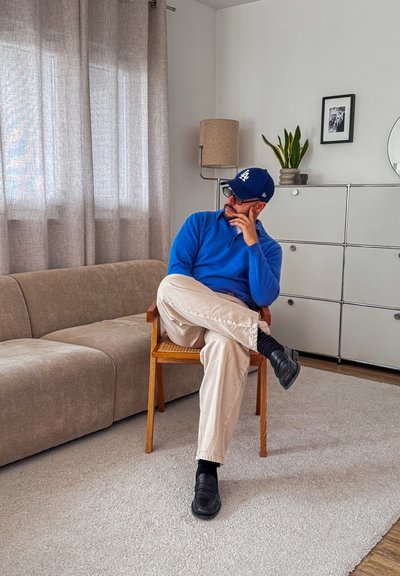 Hombre con jersey azul y gorra sentado con las piernas cruzadas en una silla de madera en una sala de estar moderna con sofá beige y armario blanco.