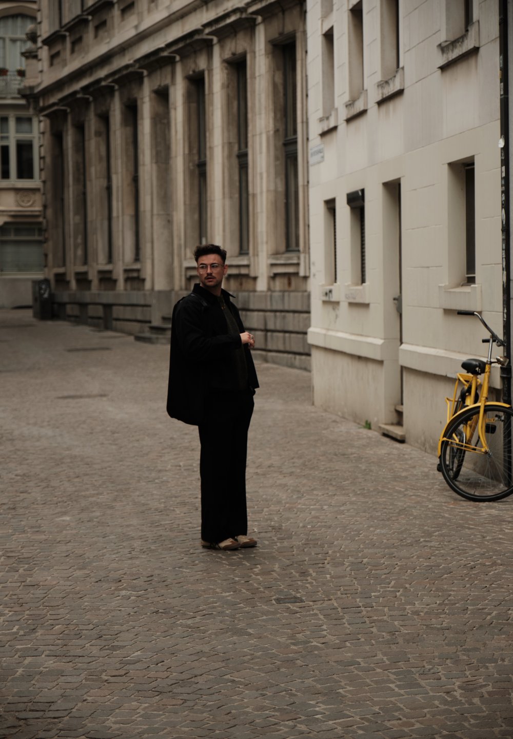 Een persoon in zwarte bovenkleding en donkere broek staat op een kasseienstraat, met een gele fiets tegen een muur leunend.