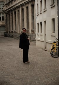 Een persoon in zwarte bovenkleding en donkere broek staat op een kasseienstraat, met een gele fiets tegen een muur leunend.