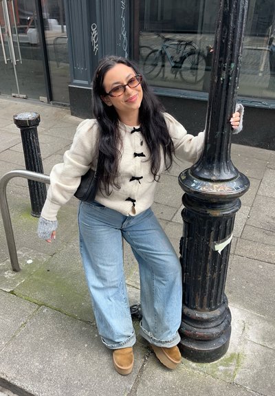 Suéter crema con botones negros, jeans de pierna ancha azules y botas de plataforma color marrón. Guantes grises sin dedos, posando junto a un poste de luz negro.