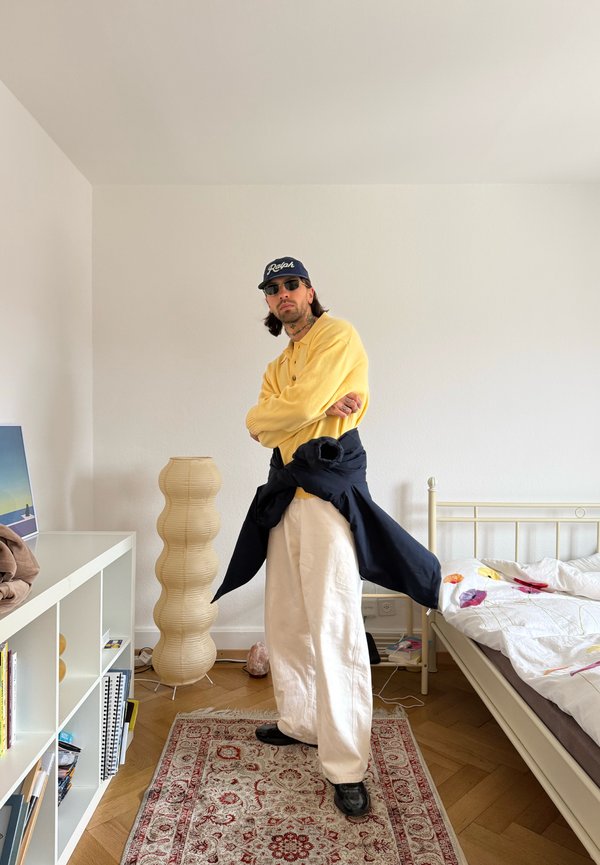 Mann trägt gelben Pullover, weiße Hose, schwarze Jacke um die Taille gebunden, schwarze Kappe und Sonnenbrille, steht in einem Schlafzimmer mit Bett und Bücherregal.