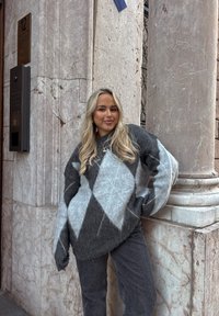 Jeune femme aux longs cheveux blonds portant un pull à losanges gris et bleu ainsi qu'un jean, debout contre un coin de bâtiment en marbre.