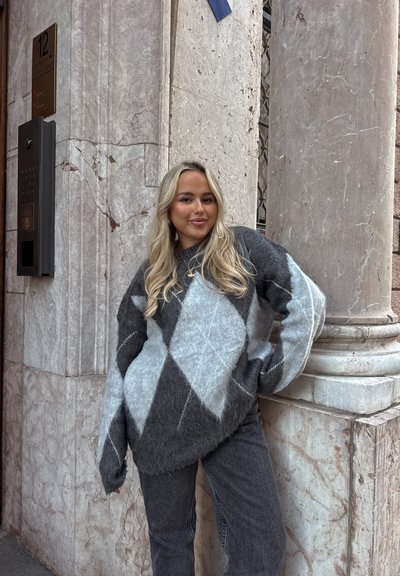 Joven con cabello largo y rubio, vistiendo un suéter de rombos gris y azul y jeans, de pie junto a la esquina de un edificio de mármol.