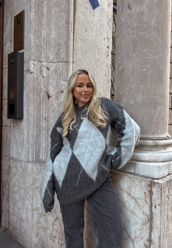 Ung kvinna med långt blondt hår, klädd i en grå och blå argyletröja och jeans, stående vid ett hörn av en byggnad i marmor.