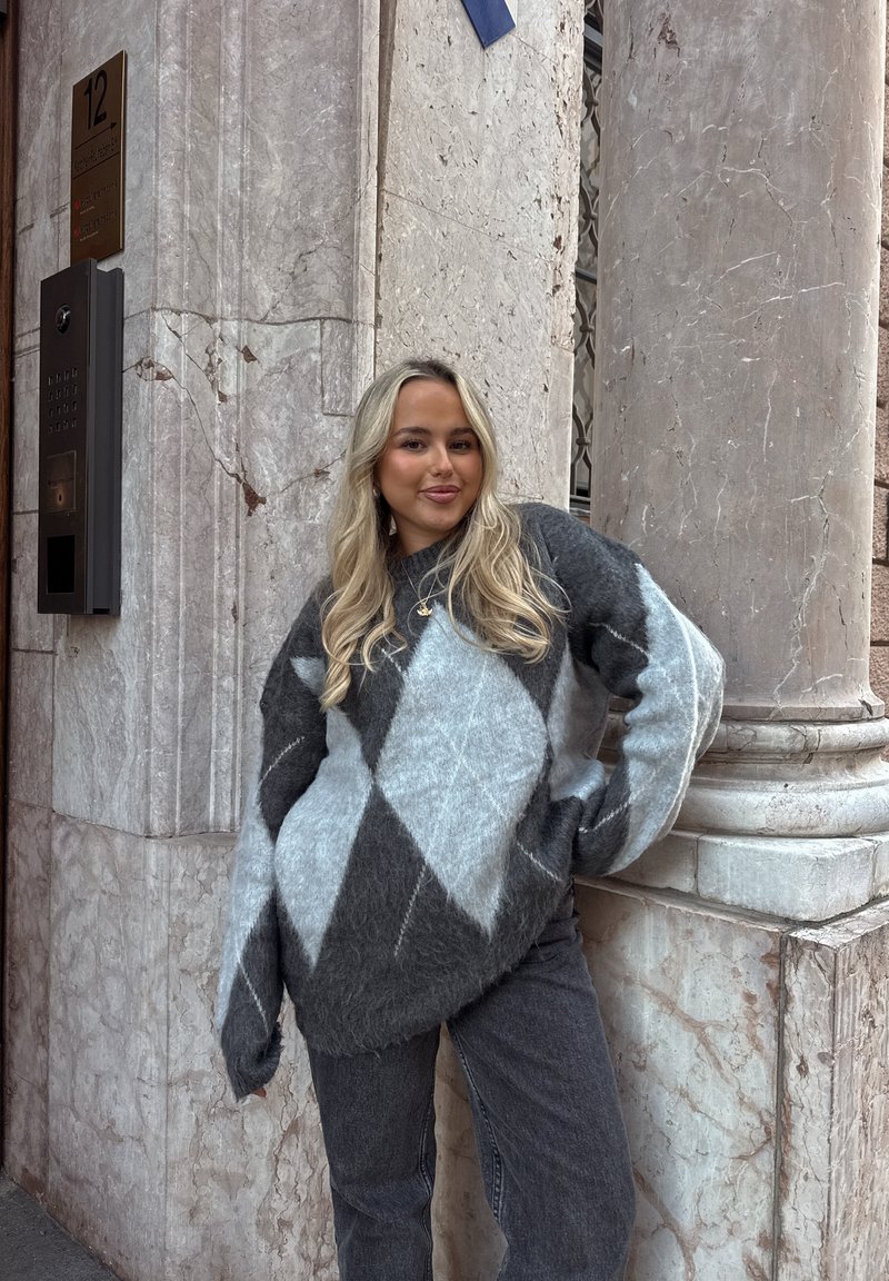 Jonge vrouw met lang blond haar, gekleed in een grijze en blauwe ruittrui en spijkerbroek, staande bij een hoek van een marmeren gebouw.