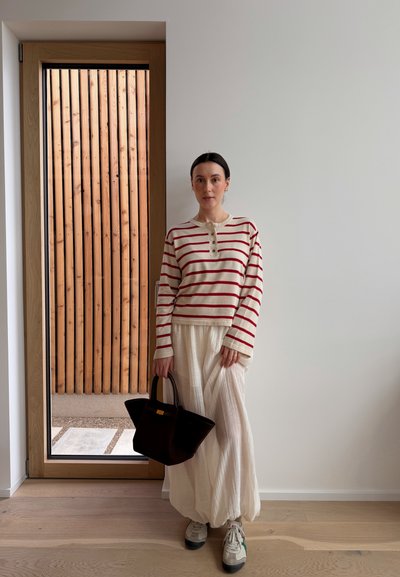 Mujer con pantalones blancos, camiseta de manga larga a rayas crema y rojo, zapatillas blancas, sosteniendo un bolso negro, de pie junto a una puerta de cristal con panel de madera en el exterior.