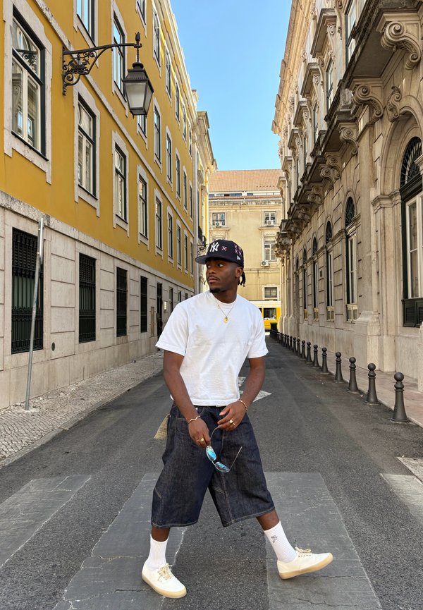 Hombre con camisa blanca y pantalones cortos oscuros caminando por una calle entre edificios amarillos y de piedra en un día soleado.