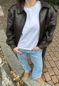 Brown leather bomber jacket with ribbed cuffs, white crewneck t-shirt, light blue wide-leg jeans, and beige slip-on shoes.