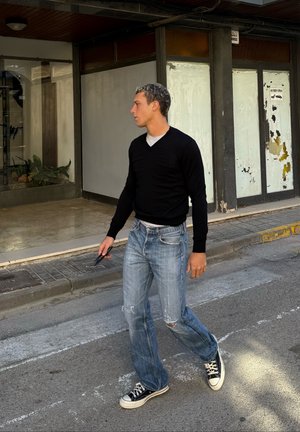 Hombre con suéter negro y jeans rotos caminando por la calle sosteniendo gafas de sol, fondo urbano con escaparate y acera.