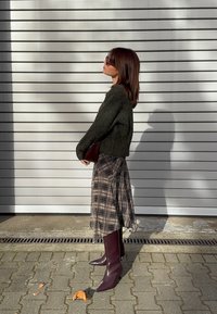 Ciemnozielony dziany sweter, midi spódnica w kratę w beżowo-brązowych odcieniach oraz bordowe, wysokie, szpiczaste kozaki za kolano; trzyma brązową kopertówkę.