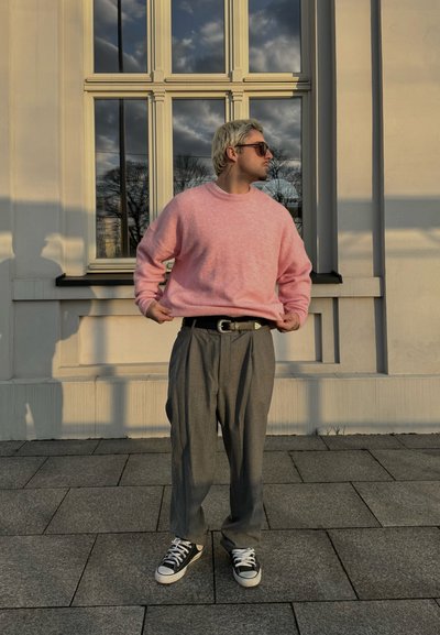Suéter de lana rosa claro, pantalones grises de cintura alta y zapatillas negras. De pie contra un edificio blanco con grandes ventanas.