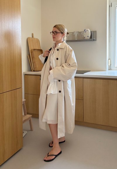 Mujer con gabardina beige, vestido blanco, gafas negras y chanclas negras de pie en una cocina moderna con armarios de madera.