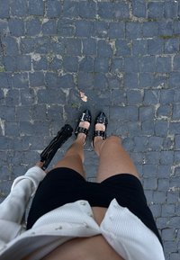 Μαύρα παπούτσια από γυαλιστερή δερματίνη με ασημένιους αγκράφες, συνδυασμένα με μια μαύρη φούστα και μια μαύρη τσάντα, σε φόντο από κυβόλιθους.