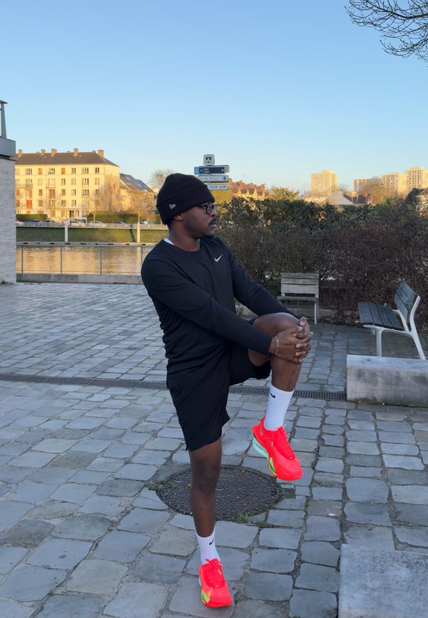 Muž v čiernom športovom oblečení a jasne červených topánkach naťahuje koleno vonku na spevnenej ploche pri rieke a lavičkách pri západe slnka.