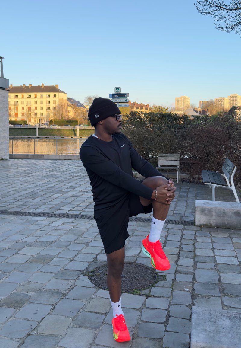 Άνδρας με μαύρη αθλητική περιβολή και φωτεινά κόκκινα παπούτσια τεντώνει το γόνατό του σε εξωτερικό χώρο σε πλακόστρωτη περιοχή κοντά σε ποτάμι και παγκάκια κατά το ηλιοβασίλεμα.