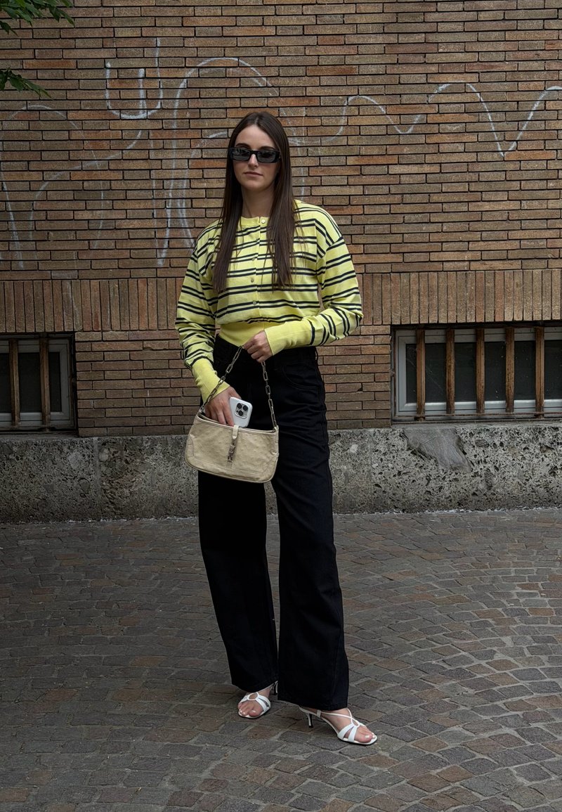Femme portant un pull rayé jaune et noir, un pantalon noir, des sandales blanches et des lunettes de soleil, tenant un sac à main beige avec un smartphone dans une rue pavée.