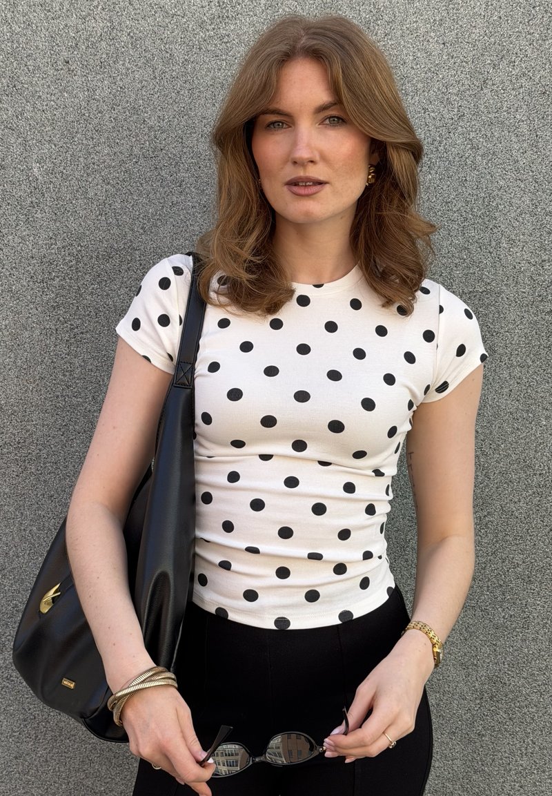 Jeune femme aux cheveux châtain clair ondulés, portant une chemise blanche à pois, un pantalon noir, des bijoux en or, tenant des lunettes de soleil noires et un sac noir.