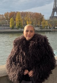 Femme aux cheveux tirés en arrière et manteau texturé foncé debout près d'une rivière en automne près de la Tour Eiffel à Paris.