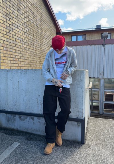 Persona con gorra roja, sudadera con capucha gris, camiseta gráfica blanca, pantalones negros y botas color canela, de pie al aire libre contra una pared de hormigón sosteniendo llaves.