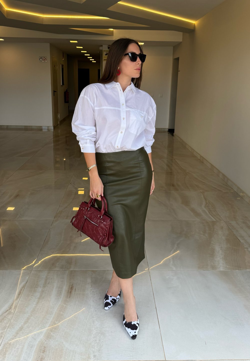 White button-up shirt, olive green leather pencil skirt, red handbag, and black-and-white patterned pointed-toe heels. Marble floor background.